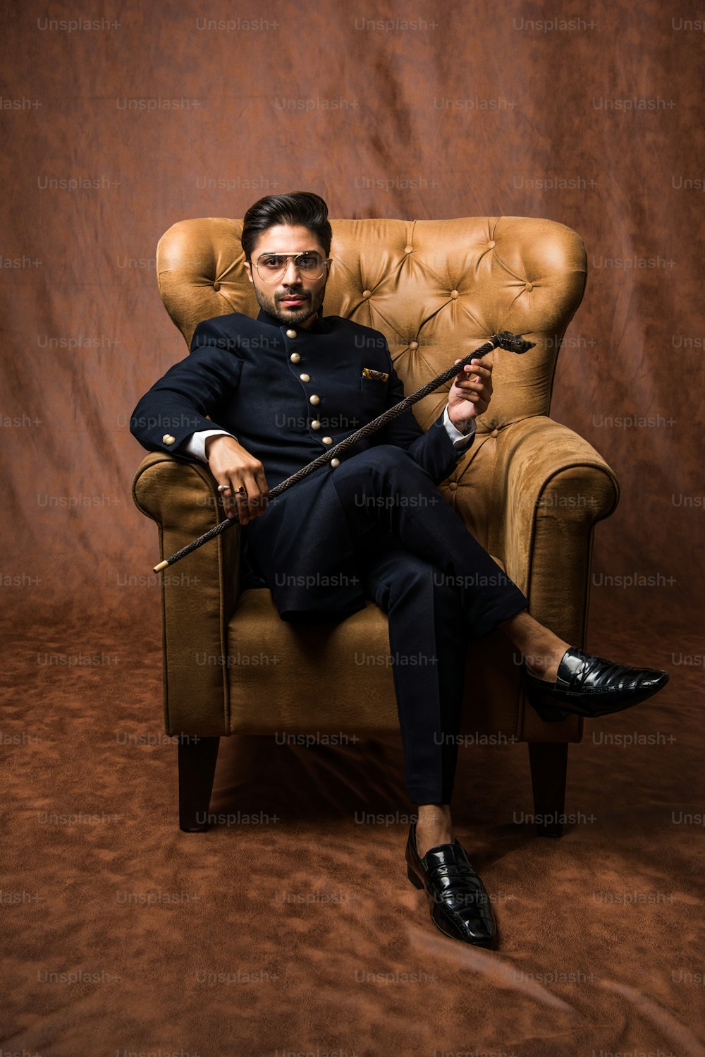 Indian bridegroom wears ethnic or traditional cloths,  Male fashion model with dark blue sherwani, Sitting on Wing Chair / sofa over brown grunge background, selective focus
