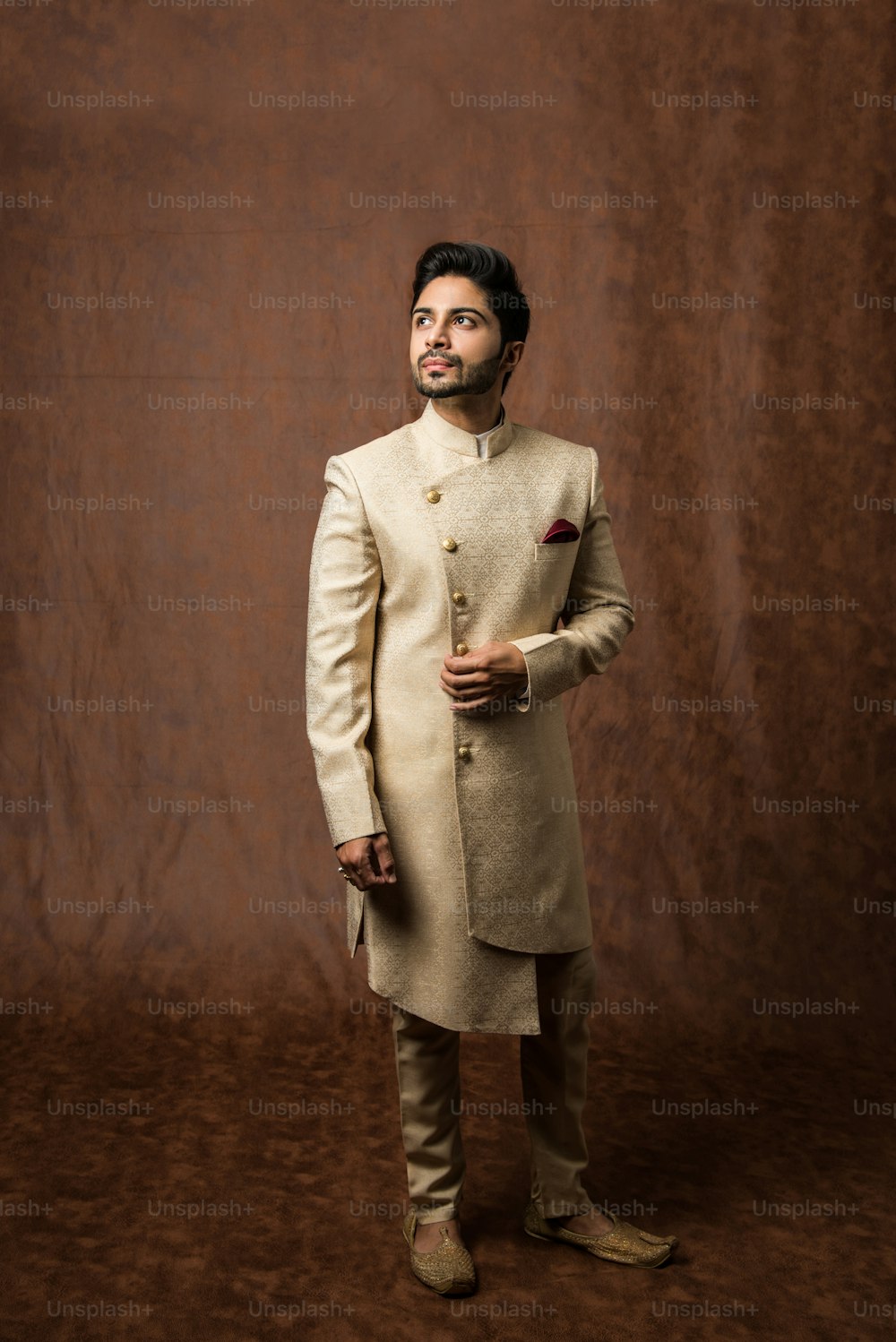 El novio indio usa kurta / telas étnicas o tradicionales.  Modelo masculino en sherwani, posando / de pie contra fondo grunge marrón, enfoque selectivo