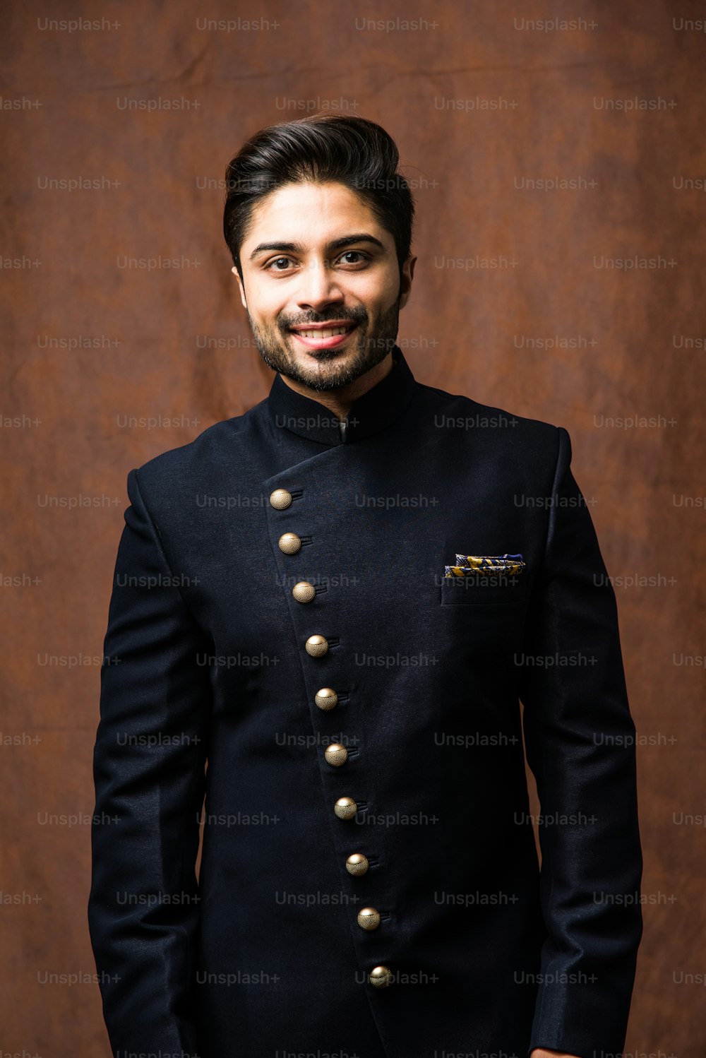 El novio indio usa telas étnicas o tradicionales, modelo de moda masculino con sherwani azul oscuro, posando / de pie contra fondo grunge marrón, enfoque selectivo