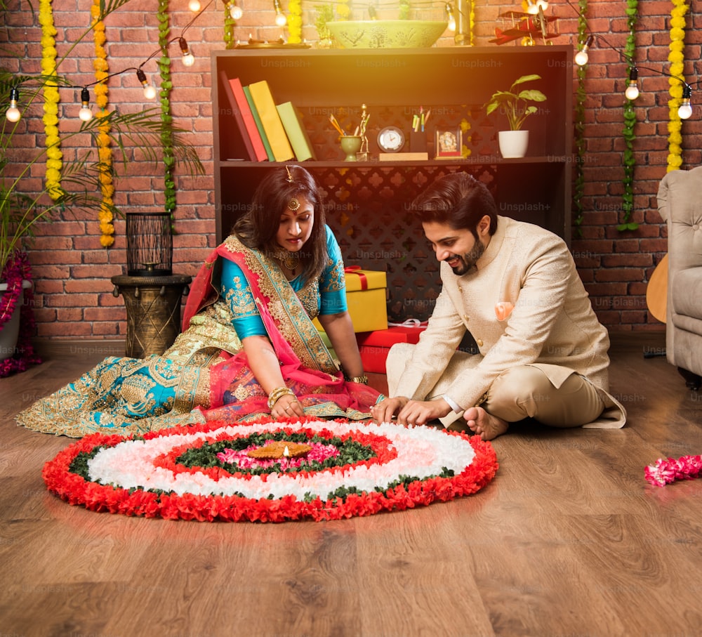 Indian couple making flower Rangoli on Diwali or Onam Festival, taking selfie or holding sweets