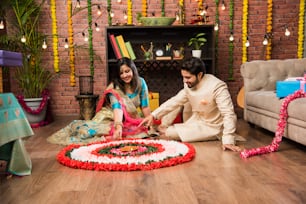 Indian couple making flower Rangoli on Diwali or Onam Festival, taking selfie or holding sweets