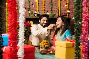 Indian smart couple eating sweet laddu on Diwali or anniversary, selective focus