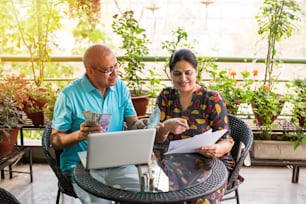 Contabilità di coppia indiana / asiatica senior, finanza domestica e controllo delle bollette con laptop, calcolatrice e denaro mentre si è seduti nel giardino di casa