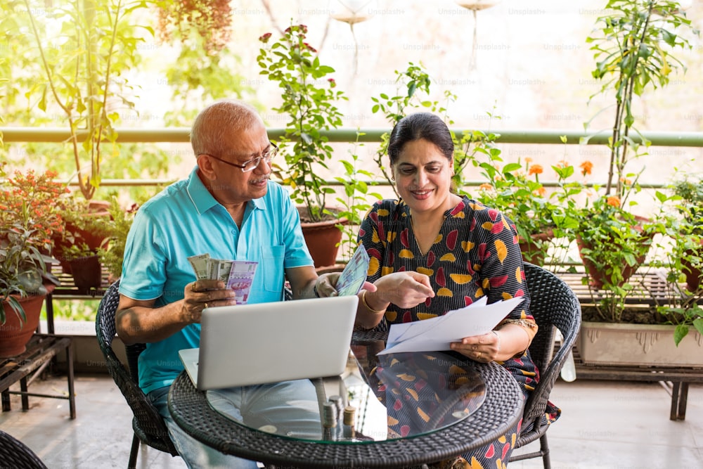 Senior Indian/asian couple accounting, doing home finance and checking bills with laptop, calculator and money while sitting at home garden