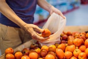Man chooses tomatoes in a supermarket without using a plastic bag. Reusable bag for buying vegetables. Zero waste concept.
