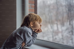 Feliz menino adorável criança sentado perto da janela e olhando para fora na neve no dia de Natal ou manhã. Criança sorridente fascinada com queda de neve e grandes flocos de neve.