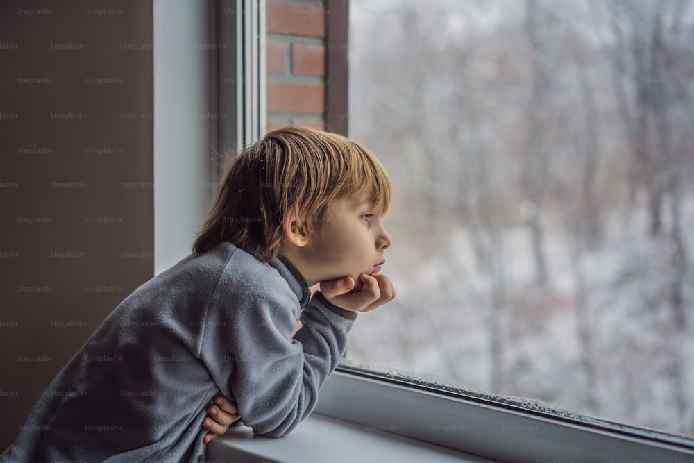 행복한 사랑스러운 아이 소년은 창가에 앉아 크리스마스 날이나 아침에 눈 위에서 밖을 내다보고 있다. 눈과 큰 눈송이에 매료된 웃는 아이.