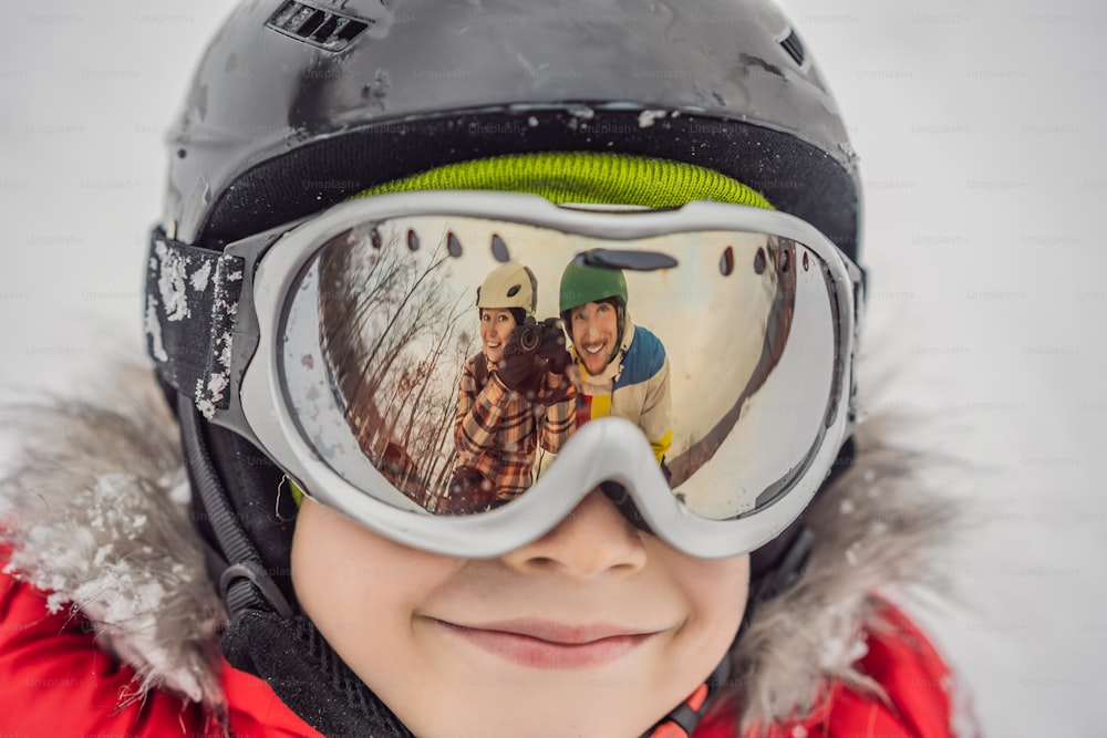 Mamãe e papai são refletidos nos óculos de esqui do menino. Mamãe e papai ensinam um menino a esquiar ou praticar snowboard.