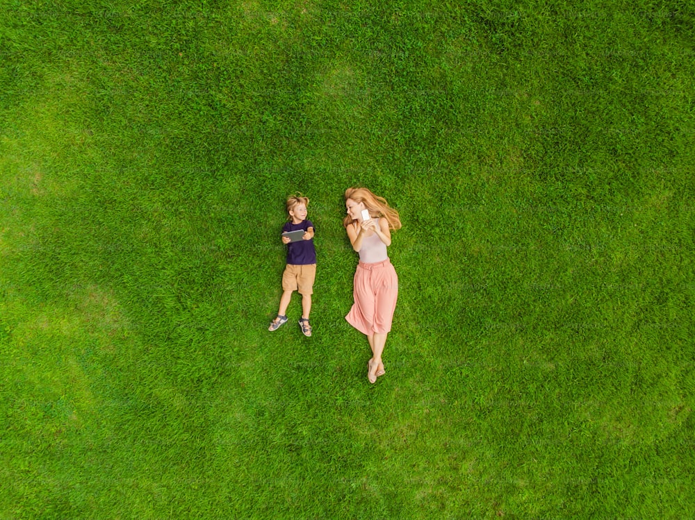 Mom and son are lying on the grass in the park. Mom looks at the phone, son looks at the tablet. Photos from the drone, quadracopter.