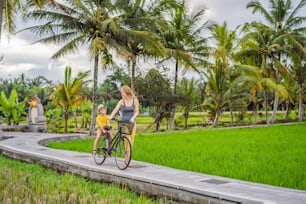 Mother and son ride a bicycle on a rice field in Ubud, Bali. Travel to Bali with kids concept.