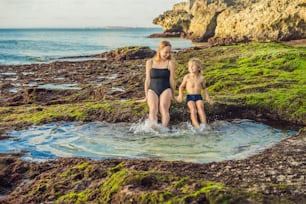 Mother and son tourists on Pantai Tegal Wangi Beach sitting in a bath of sea water, Bali Island, Indonesia. Bali Travel Concept. Traveling with children concept. Kids friendly places.