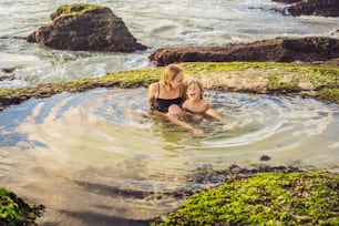 Mother and son tourists on Pantai Tegal Wangi Beach sitting in a bath of sea water, Bali Island, Indonesia. Bali Travel Concept. Traveling with children concept. Kids friendly places.