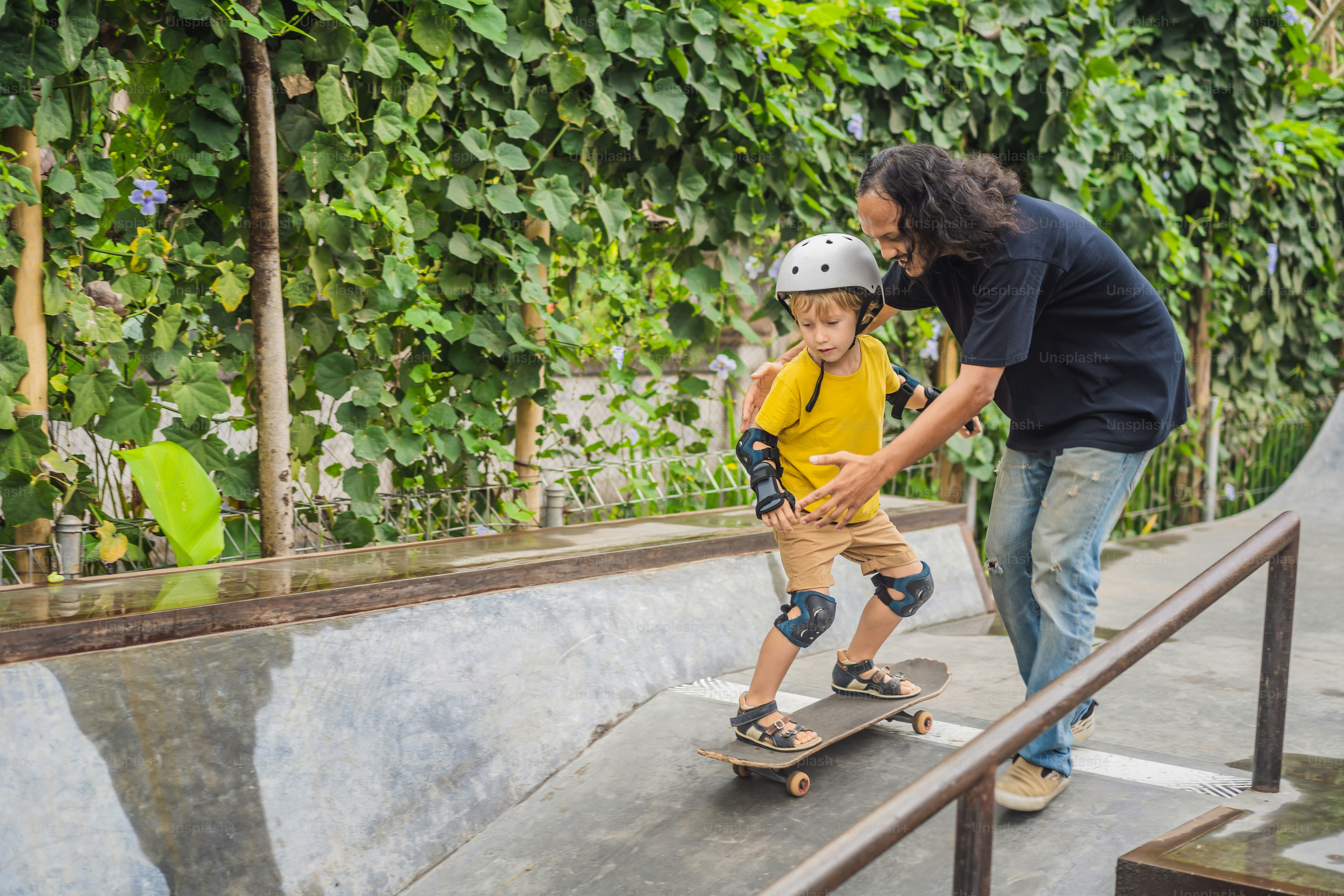 Athletic boy learns to skateboard with a trainer in a skate park ...