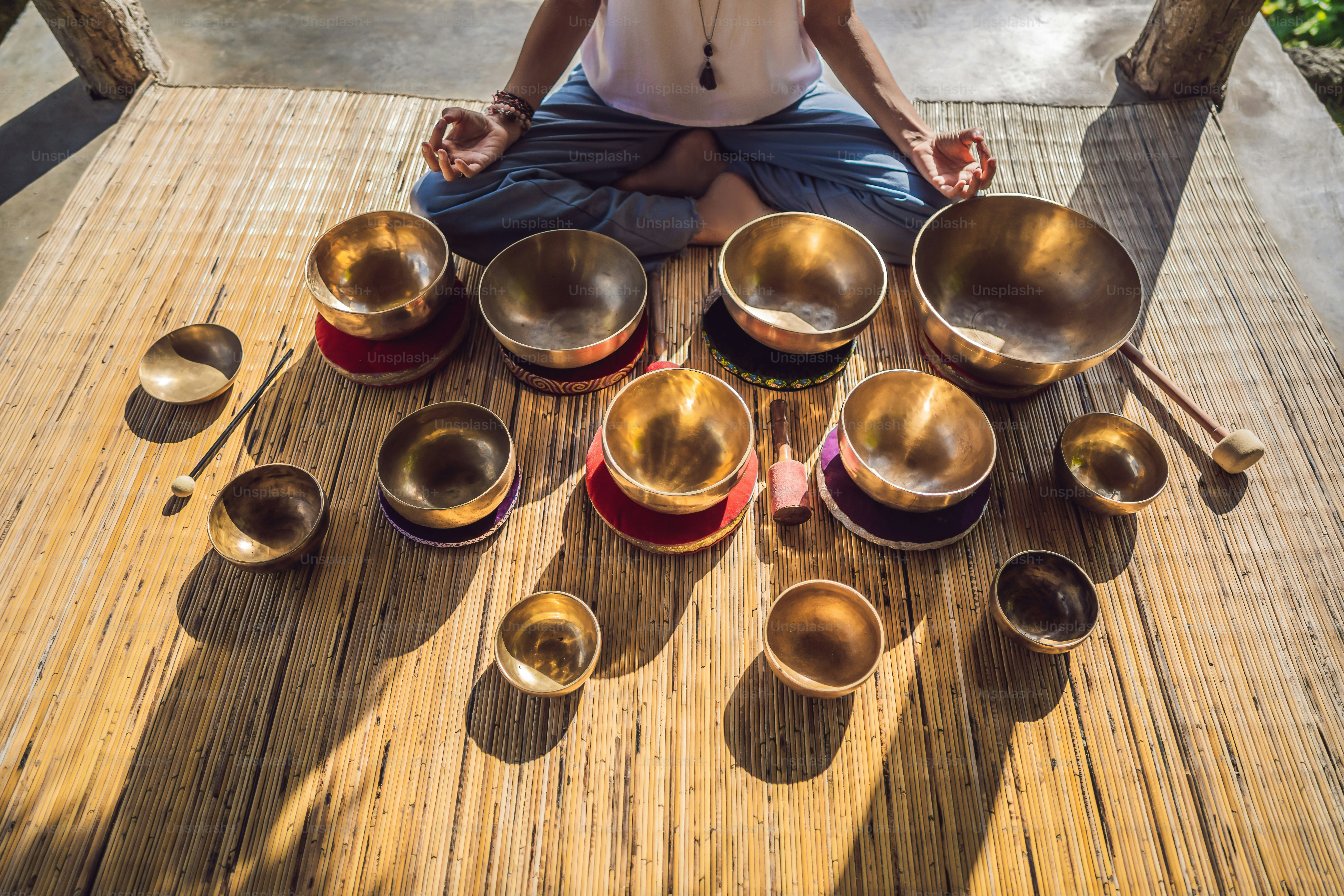 Woman playing on Tibetan singing bowl while sitting on yoga mat against a waterfall. Vintage tonned. Beautiful girl with mala beads meditating.