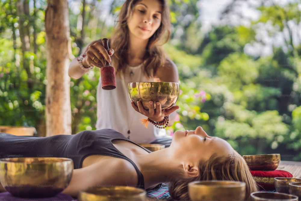 Cuenco cantor de cobre de Buda de Nepal en el salón de spa. Joven hermosa mujer haciendo terapia de masaje cantando cuencos en el Spa contra una cascada. Terapia de sonido, recreación, meditación, estilo de vida saludable y concepto de cuidado corporal.