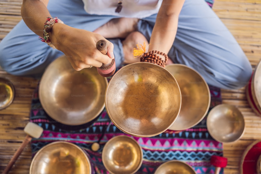 Woman playing on Tibetan singing bowl while sitting on yoga mat against a waterfall. Vintage tonned. Beautiful girl with mala beads meditating.