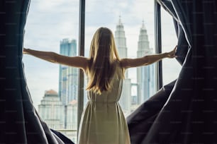 Young woman opens the window curtains and looks at the skyscrapers in the big city.