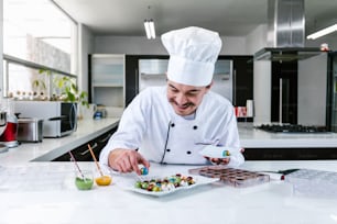 junger lateinamerikanischer Mann Chocolatier mit Kochmütze, der mit mexikanischen Pralinen, Süßigkeiten auf dem Teller in einer Großküche in Mexiko, Lateinamerika, steht