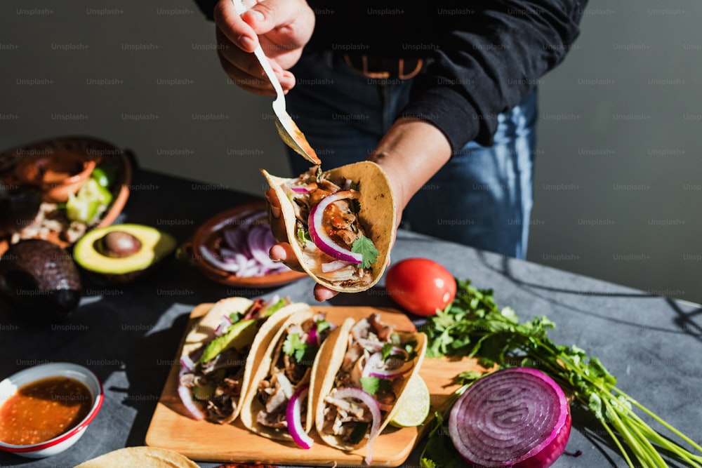 latin woman hands preparing mexican tacos with pork carnitas, avocado, onion, cilantro, and red sauce in Mexico