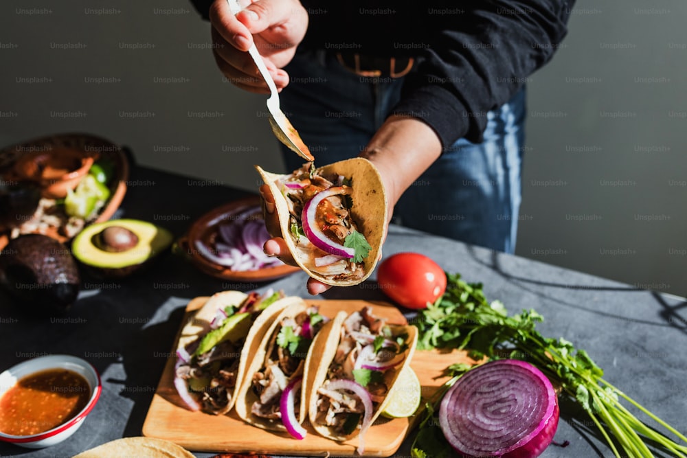 lateinamerikanische Frauenhände, die mexikanische Tacos mit Schweinefleisch-Carnitas, Avocado, Zwiebeln, Koriander und roter Soße in Mexiko zubereiten