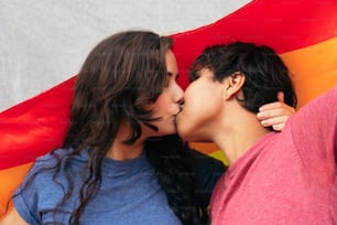 Ein junges Mädchenpaar, das sich an einem sonnigen Tag in einem Park mit einer Gay-Pride-Flagge küsst, den Pride Day feiert und sich verliebt.