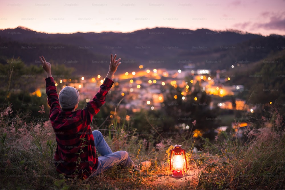 Jovem sentindo-se livre e motivado no topo de uma montanha com uma lanterna de querosene acesa, observando as luzes da cidade à noite.