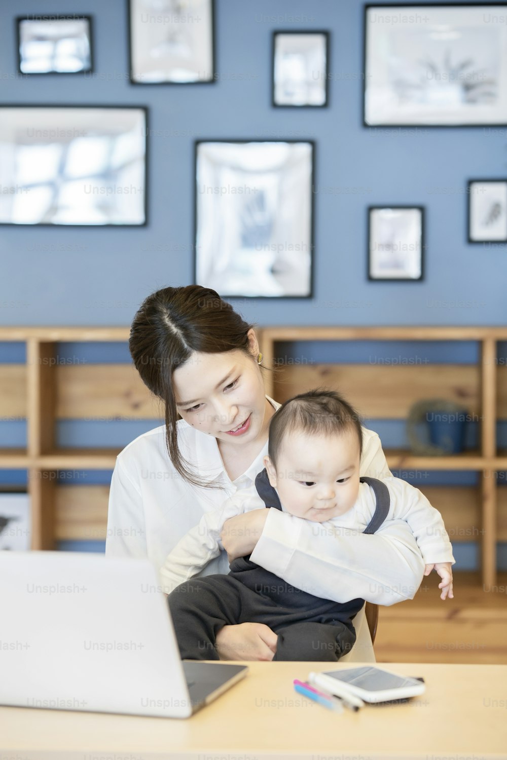 Asian woman holding a baby and operating a laptop indoors