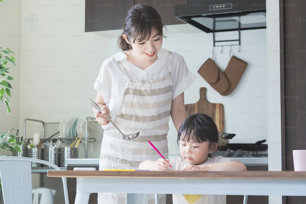 A girl drawing in the kitchen and a mother watching over her while cooking