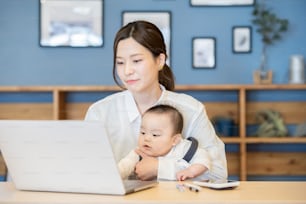Asian woman holding a baby and operating a laptop indoors