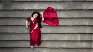 Portrait of beautiful Indian girl in heritage stepwell wearing traditional Indian red saree, gold jewellery and bangles. Maa Durga agomoni shoot concept. Traditional woman on stairs with poses