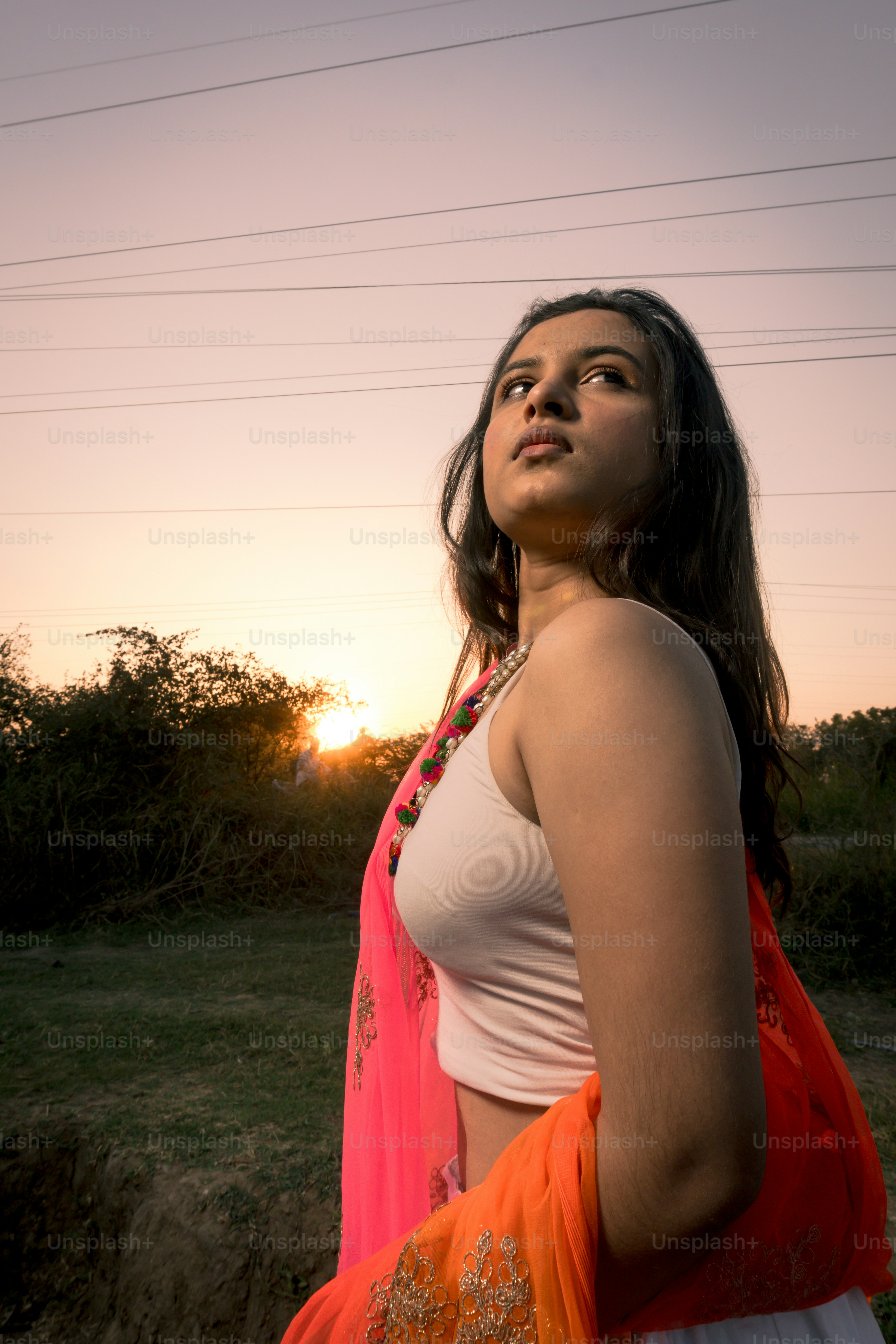 A woman in a white top and orange sari looking toward the low sun