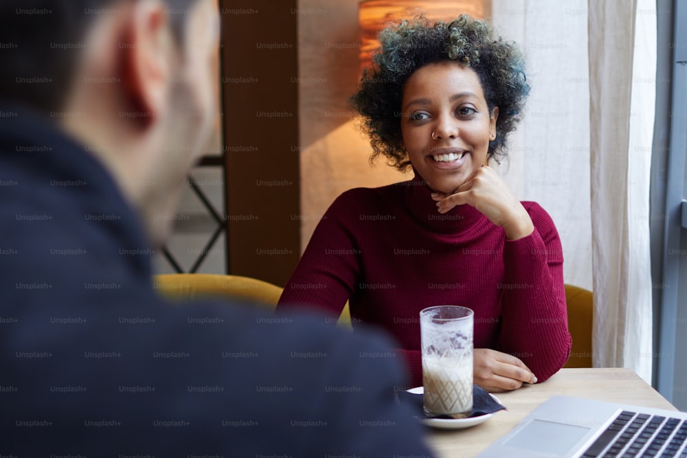 Innenfoto einer jungen schönen afroamerikanischen Dame, die hinter dem Rücken ihres Freundes am Cafétisch sitzt, ihr Kinn auf die Hand legt, glücklich lächelt und Liebe, Hingabe und Freude zeigt.