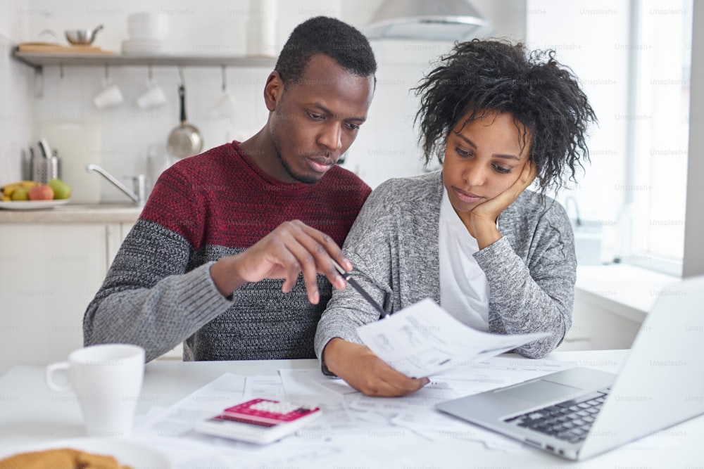 Jeune femme et mari afro-américain assis à la maison avec un ordinateur portable, une calculatrice et des papiers faisant de la paperasse ensemble, analysant les dépenses, planifiant le budget familial et calculant les factures, n’ayant pas d’argent