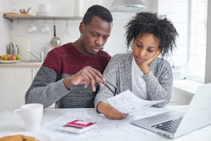 Junge afroamerikanische Ehefrau und Ehemann sitzen zu Hause mit Laptop, Taschenrechner und Papieren, erledigen gemeinsam Papierkram, analysieren Ausgaben, planen Familienbudget und berechnen Rechnungen, haben kein Geld