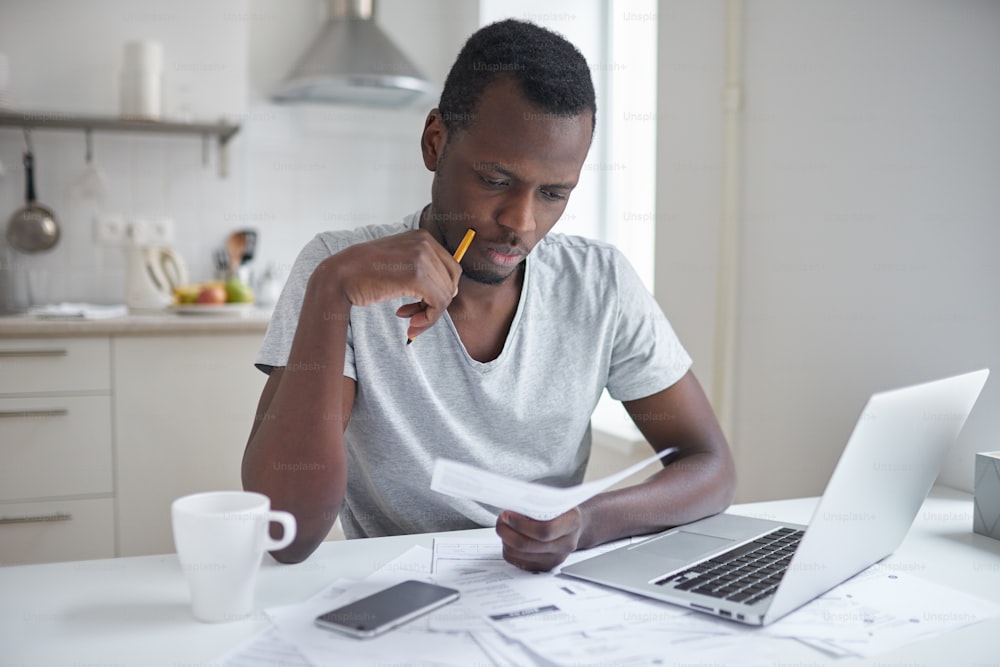 Homem afro-americano estressado trabalhando através de papelada, calculando despesas, tentando economizar algum dinheiro, gerenciando finanças, sentando-se à mesa da cozinha com laptop, tentando fazer plano de finanças