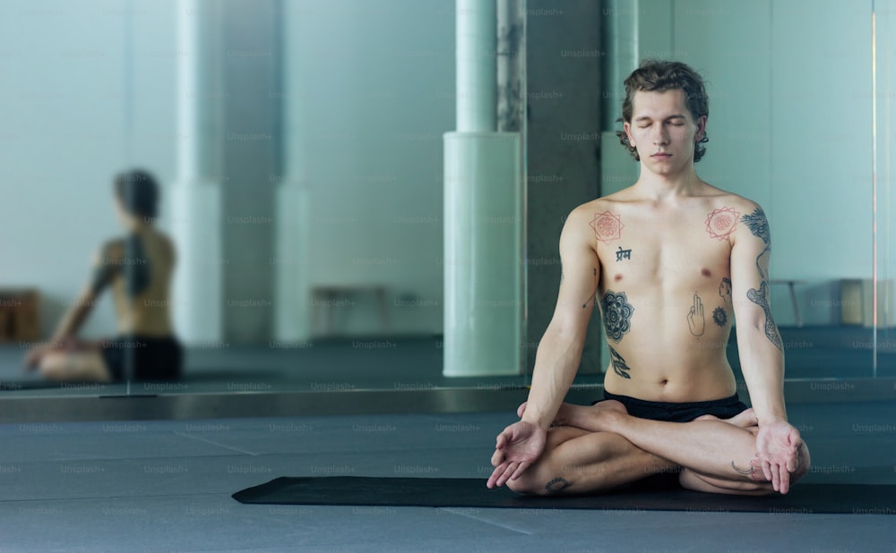 Handsome tattooed man practicing yoga. Young male teacher meditating in lotus pose with closed eyes in class on black mat. Yogi master concept
