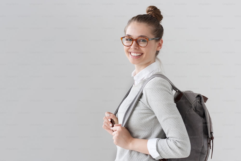 Good-looking European female on right side of photo isolated on gray background pulling forward straps of backpack with expression of satisfaction and happiness, looking hopeful and energetic.