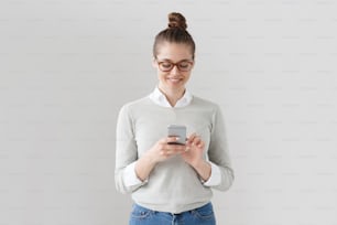 Indoor shot of young attractive European female in gray pullover, white shirt and brown-rimmed glasses isolated on gray background with smartphone in hand, touching screen with finger to type message.