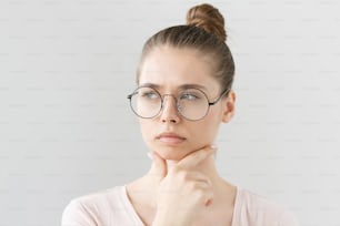 Headshot of good-looking young female in glasses isolated on gray background with head turned leftwards, scratching chin with fingers in deep thinking with uncertain expression and doubt on face.