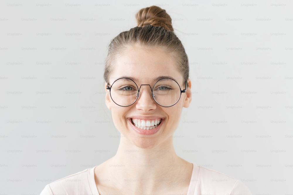 Innenporträt einer jungen schönen Frau, isoliert auf grauem Hintergrund, lächelnd, blinzelnd hinter einer großen runden Brille mit Ausdruck von vollem Inhalt, Zufriedenheit und Glück mit der aktuellen Situation.