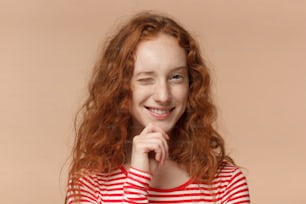 Headshot in studio di una ragazza adolescente dai capelli rossi di bell'aspetto isolata su sfondo pesca che sembra intraprendente ed entusiasta, ammiccante amichevole come se invitasse all'avventura o raccomandasse un buon beneficio