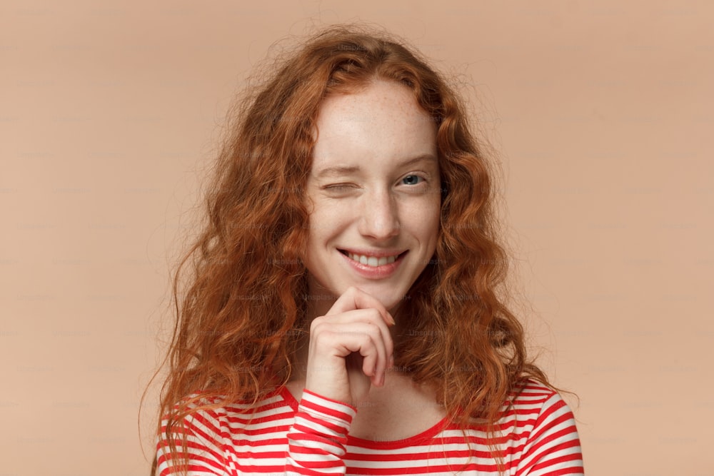 桃の背景に見栄えの良い赤毛の10代の女の子のスタジオヘッドショットは、進取的で熱狂的に見え、冒険に招待したり、良い利益を勧めたりするかのように友好的にウィンクします