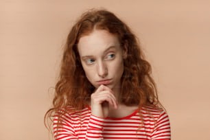Closeup headshot of beautiful redhead woman isolated on peach background looking sidewards feeling distrust, hesitation, doubting given facts, uneasy to communicate and cooperate, showing discontent