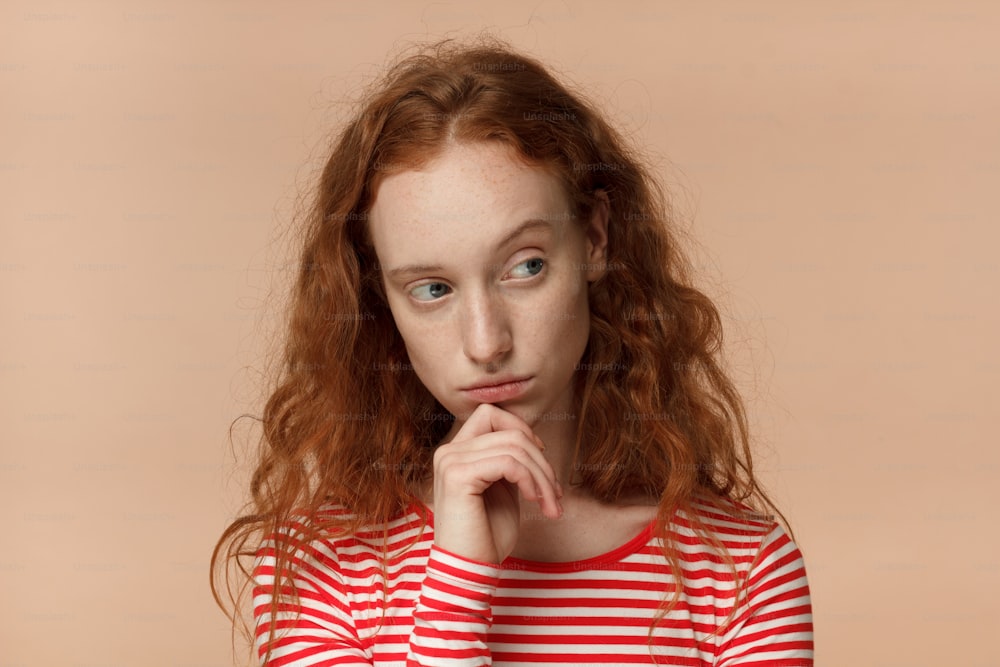 Closeup headshot of beautiful redhead woman isolated on peach background looking sidewards feeling distrust, hesitation, doubting given facts, uneasy to communicate and cooperate, showing discontent
