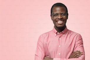 Bel homme afro-américain souriant en chemise formelle isolé sur fond rose debout les bras croisés