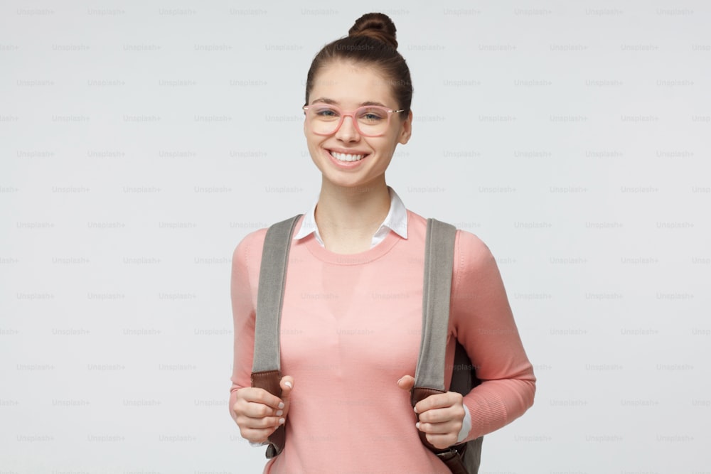 Portrait of happy young woman in casual clothes and trendy pink eyeglasses standing isolated on grey background with backpack on her back