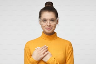 Lovely woman in yellow polo neck sweater having charming smile and friendly expression holding hands on her heart, showing her love and sympathy.