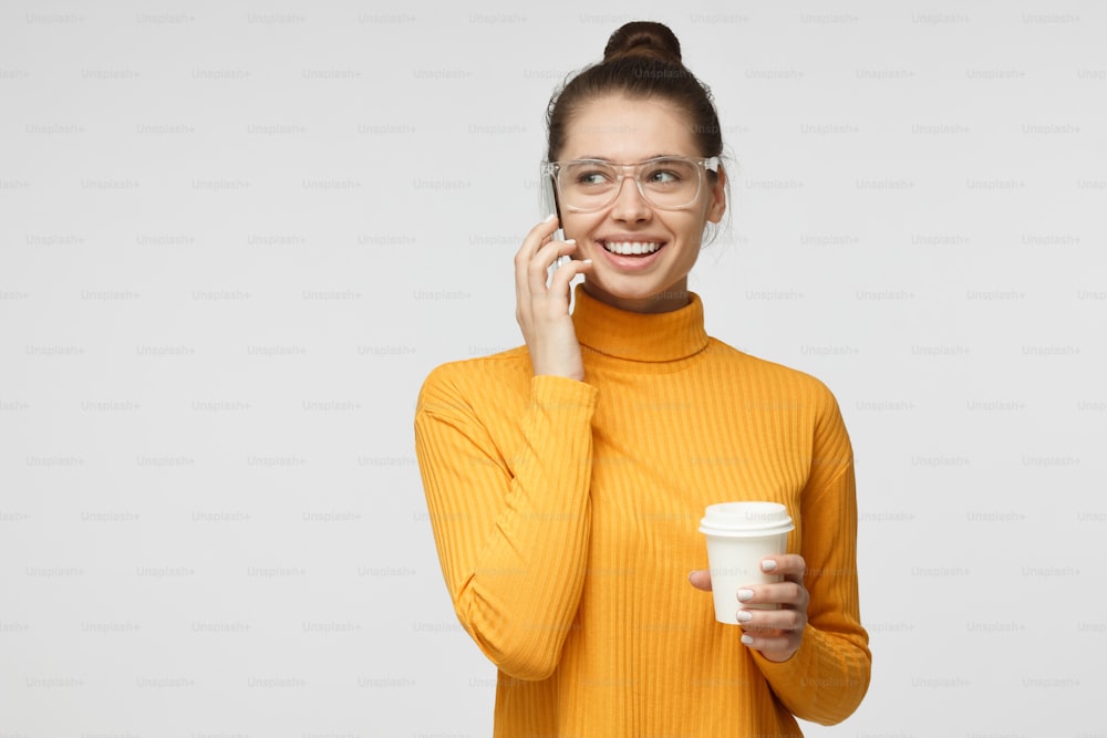 Horizontal picture of young attractive female wearing casual clothes holding smartphone next to ear and cup of coffee to go, looking sidewards while talking with friend with smile