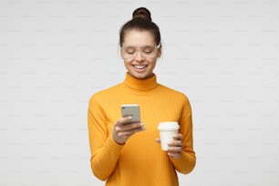 Imagem interna da jovem mulher europeia de boa aparência isolada no fundo cinza segurando o telefone na mão, olhando para a tela sorrindo enquanto navega ou lê a mensagem