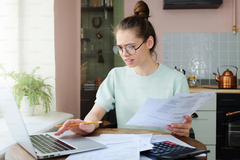 Horizontal shot of European Caucasian female wearing T-shirt and glasses sitting at table with laptop and bills trying to check possible mistakes in calculations with concerned and worried face